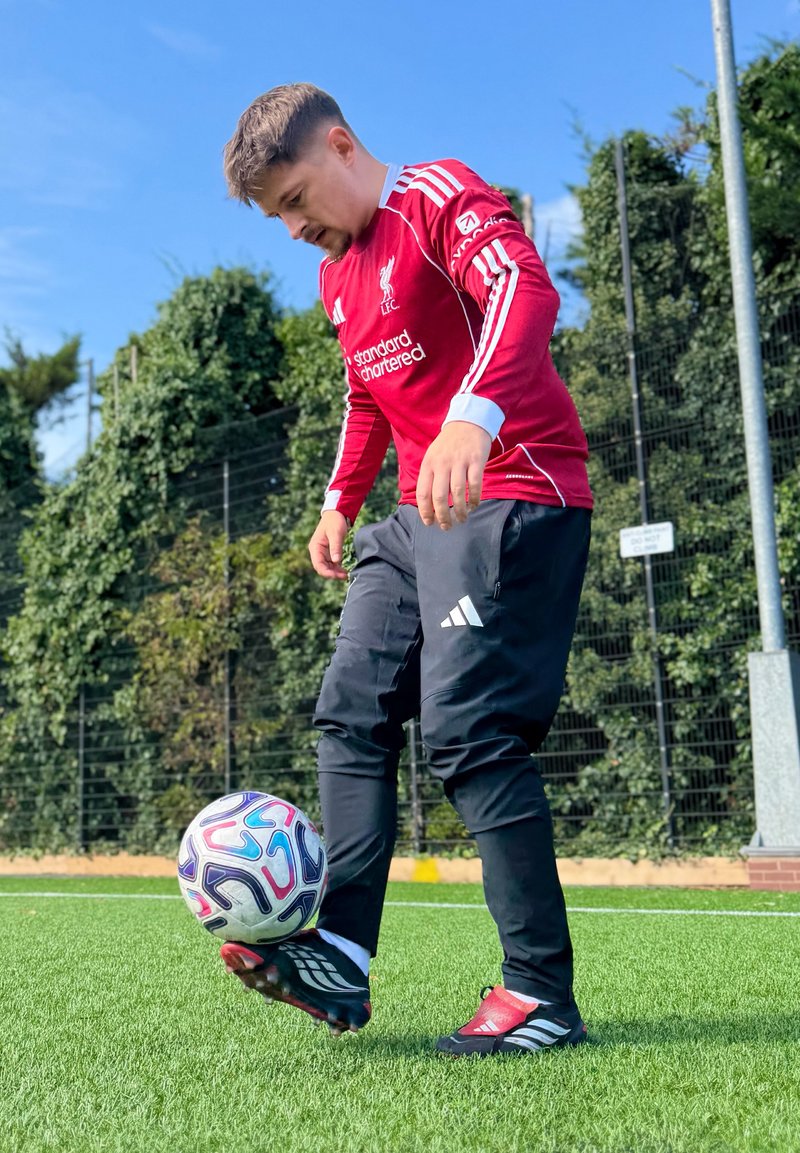 Homme en maillot rouge de Liverpool et pantalon Adidas noir contrôlant un ballon de football sur un terrain en herbe avec végétation et clôture en arrière-plan.