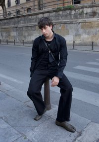 Fekete gombos ing, laza szabásban, fekete farmernadrággal párosítva. Kiegészítők között szerepel egy ezüst nyaklánc. A modell egy utcai oszlopra ül.