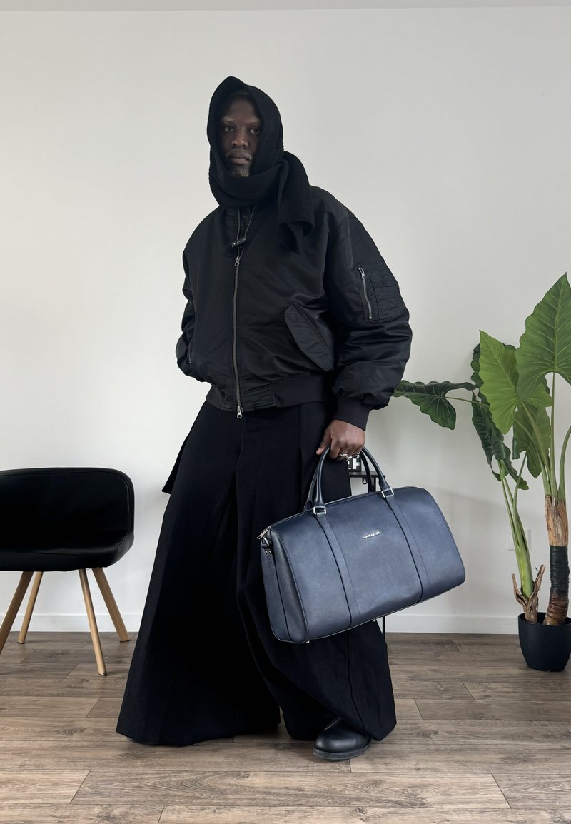Black bomber jacket, long black skirt, and black ankle boots. Holding a blue leather bag with two handles and a textured surface.