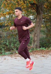 Chemise et pantalon de sport bordeaux avec des accents réfléchissants. Baskets orange vif et rose avec une semelle bleu clair. Coureur sur un chemin pavé.