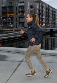 Egy futó tengerészkék széldzsekiben, barna mintás leggingsben és zöldes-szürke, fényvisszaverő részletekkel ellátott futócipőben, betonúton kocog.