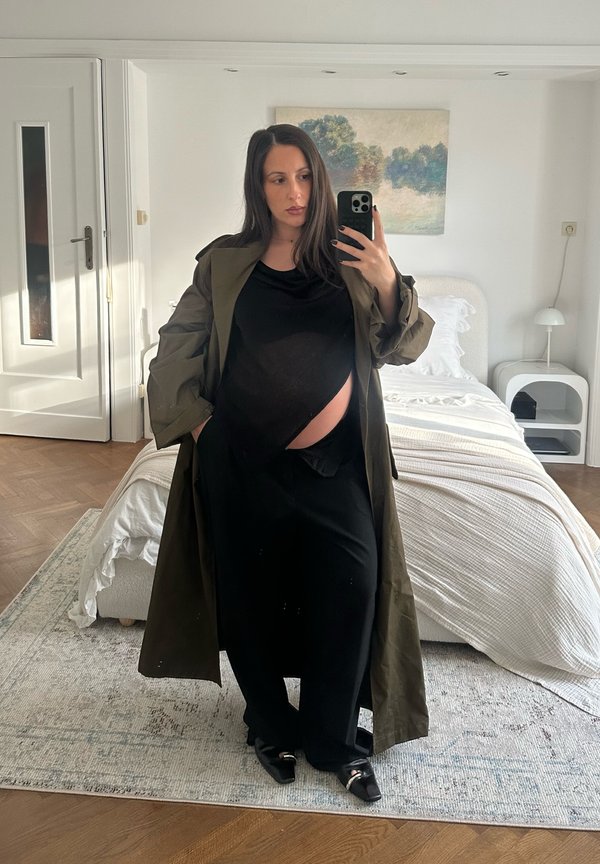 Mujer embarazada con vestido negro y abrigo verde tomando una selfie frente al espejo en un dormitorio luminoso con cama, mesita de noche y cuadro.