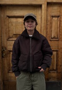 Bruine bomberjas met een zachte kraag, ritssluiting aan de voorkant en twee grote zakken. Gecombineerd met losse groene broek. Houten achtergrond.