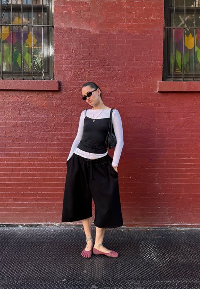 Top negro ajustado superpuesto sobre una camisa blanca de manga larga, combinado con pantalones cortos anchos negros y zapatos morados tipo slip-on. Bolso bandolera negro.