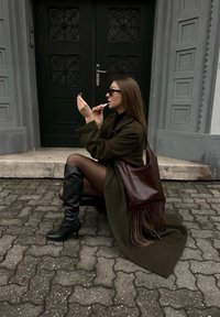 Femme en manteau sombre et bottes assise sur des marches en pavé, appliquant du rouge à lèvres tout en tenant un petit miroir devant une grande porte sombre.