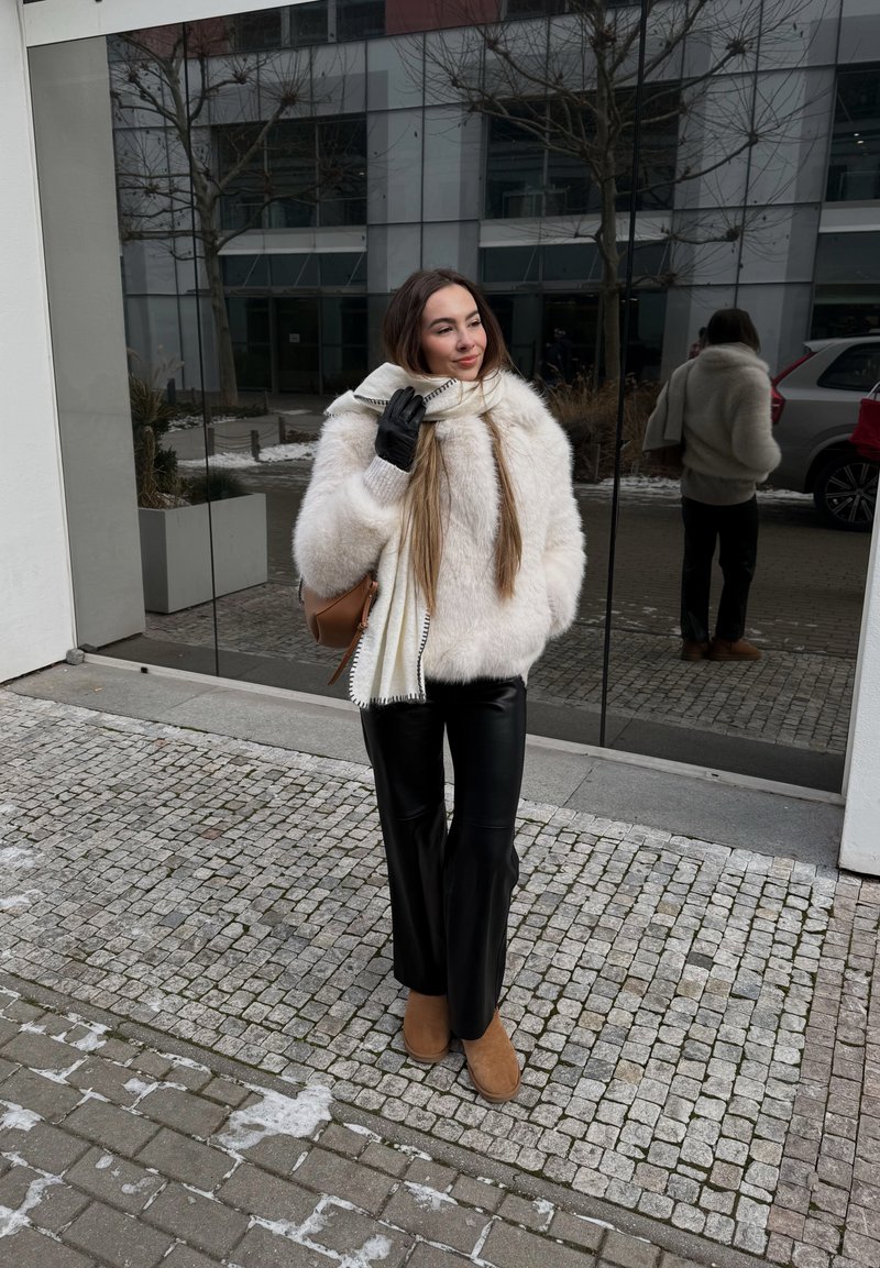 Femme portant une veste en fourrure blanche, un pantalon en cuir noir, des bottes marron et une écharpe crème, debout sur un trottoir pavé près d'un mur en verre réfléchissant.