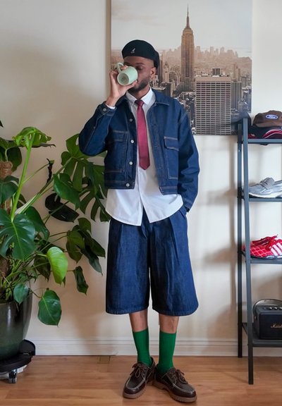 Chaqueta de denim con detalles de remaches, camisa blanca, corbata roja, pantalones cortos de denim anchos, calcetines verdes, zapatos marrones y una taza verde. Ambiente interior con plantas.