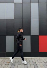 Femme athlétique portant une veste noire et un legging, chaussures de course sur un mur gris texturé avec des panneaux noirs et rouges.