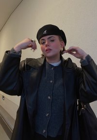 Μαύρο δερμάτινο μπουφάν πάνω από ένα γκρι πουλόβερ με κουμπιά, φορώντας ένα μαύρο μπερέ και μεγάλα χρυσά σκουλαρίκια, με μια υφασμένη τοιχοποιία από πίσω.