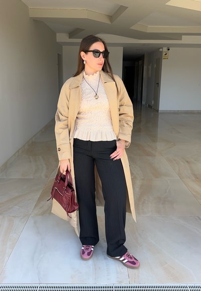 Mujer que lleva un abrigo beige tipo gabardina, blusa blanca con textura, pantalones negros de rayas finas, zapatillas rosas, gafas de sol y sostiene un bolso granate en interiores.