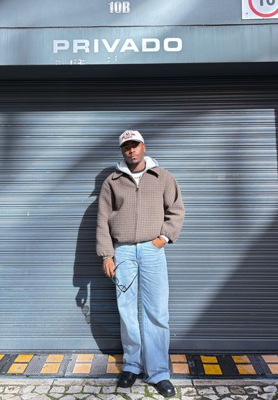 Hombre con gorra, chaqueta a cuadros y vaqueros, de pie con una mano en el bolsillo y sosteniendo unas gafas de sol, frente a una persiana metálica cerrada con un cartel que dice "Privado".