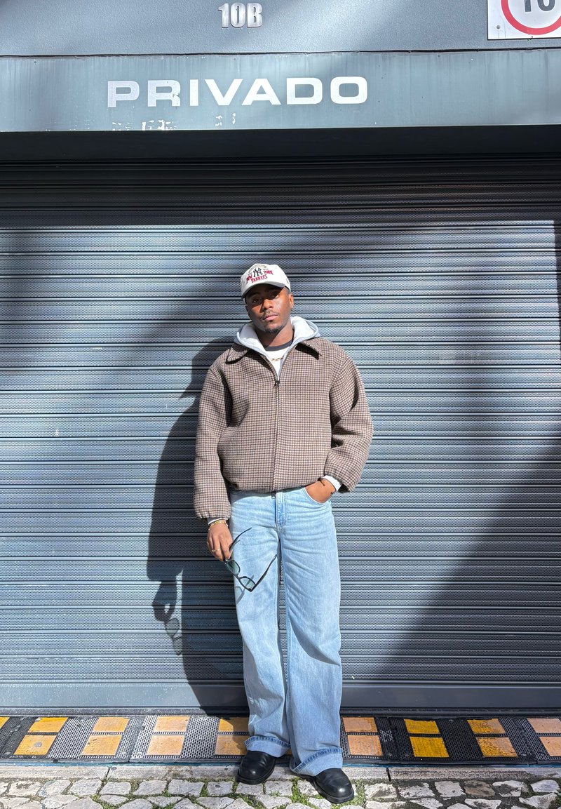 Mees, kellel on peas nokamüts, ruuduline jakk ja teksad, seisab ühe käega püksitaskus ja hoiab teises käes päikeseprille, metallist sulgemiskardina ees, millel on silt "Privado".