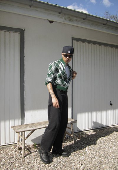 Cardigan a cuadros verdes y blancos sobre una camisa blanca, pantalones anchos negros y zapatos negros; el fondo presenta una pared blanca y un banco.