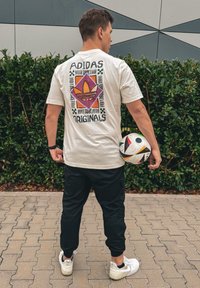 Λευκή μπλούζα με πολύχρωμο σχέδιο που περιλαμβάνει το λογότυπο της Adidas στην πλάτη; σε συνδυασμό με μαύρο παντελόν ι και κρατώντας μια μοτίβο ποδοσφαιρική μπάλα.