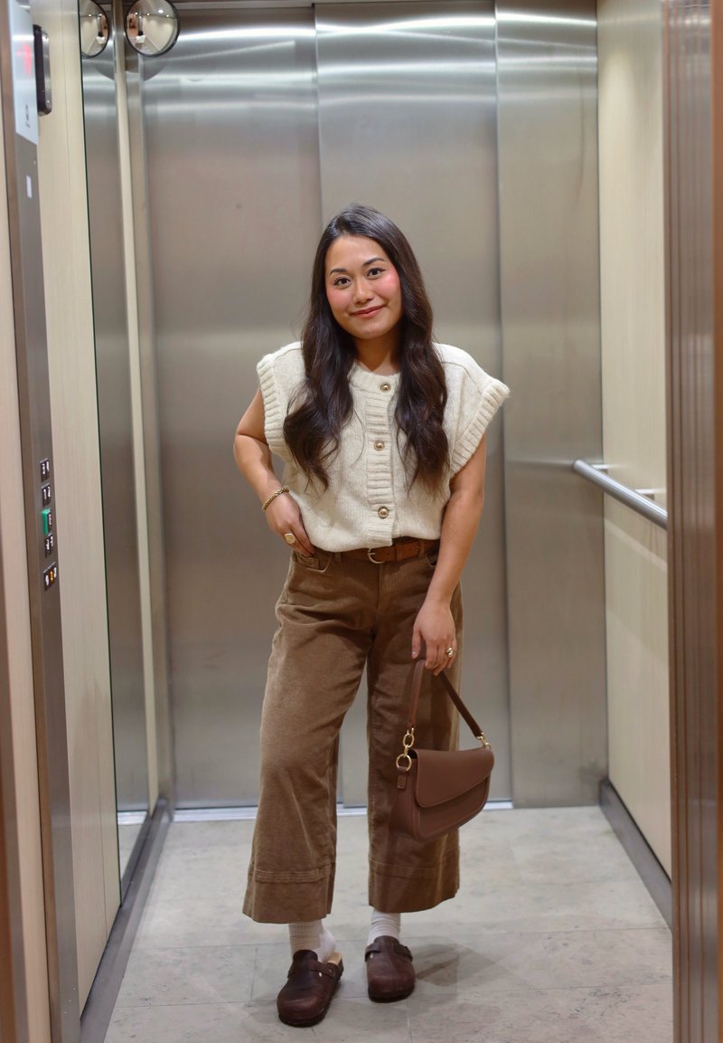 Cremefarvet striktop med ribbet struktur og knapper; brune viddeben bukser i fløjl; brun håndtaske; brune træsko; baggrund med elevator.