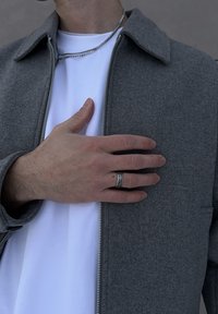 Veste grise zippée avec col portée sur un T-shirt blanc. La main posée sur la poitrine porte une bague texturée, dévoilant le ton de peau et les détails de la main.
