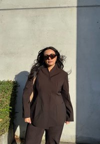 Brun skræddersyet jakke med spidse kraver, parret med matchende bukser. Tekstureret stof og lange ærmer. Overdimensionerede skildpaddemønstrede solbriller iført.