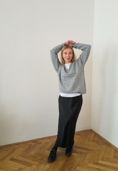 Suéter gris de punto, camisa blanca debajo, falda larga negra y zapatos negros. Entorno interior con suelo de madera clara y paredes simples.