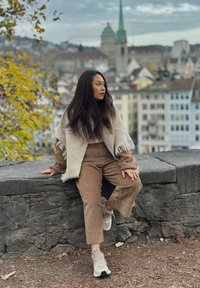Poncho beige tissé avec des franges, pantalon en velours côtelé marron et baskets claires. En arrière-plan, un paysage urbain avec des bâtiments historiques.
