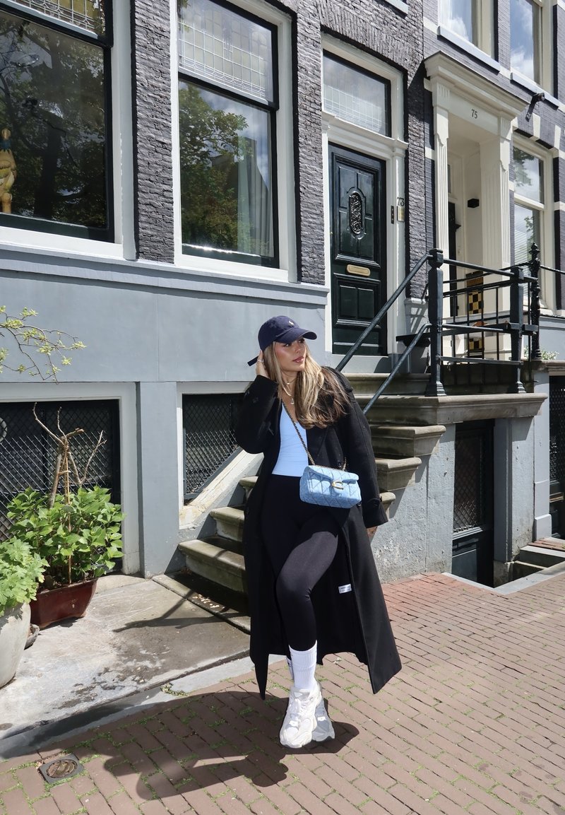 Vrouw met zwarte jas, zwarte pet, wit topje en sneakers, staand op een bakstenen stoep nabij de trap van een grijs gebouw met zwarte deur en ramen.