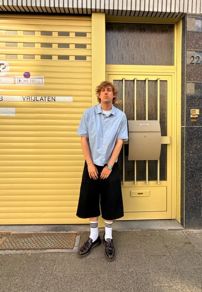 Camisa de manga corta a rayas en azul claro, pantalones cortos negros hasta la rodilla, calcetines blancos con rayas negras y zapatos negros. Fondo amarillo con un buzón.