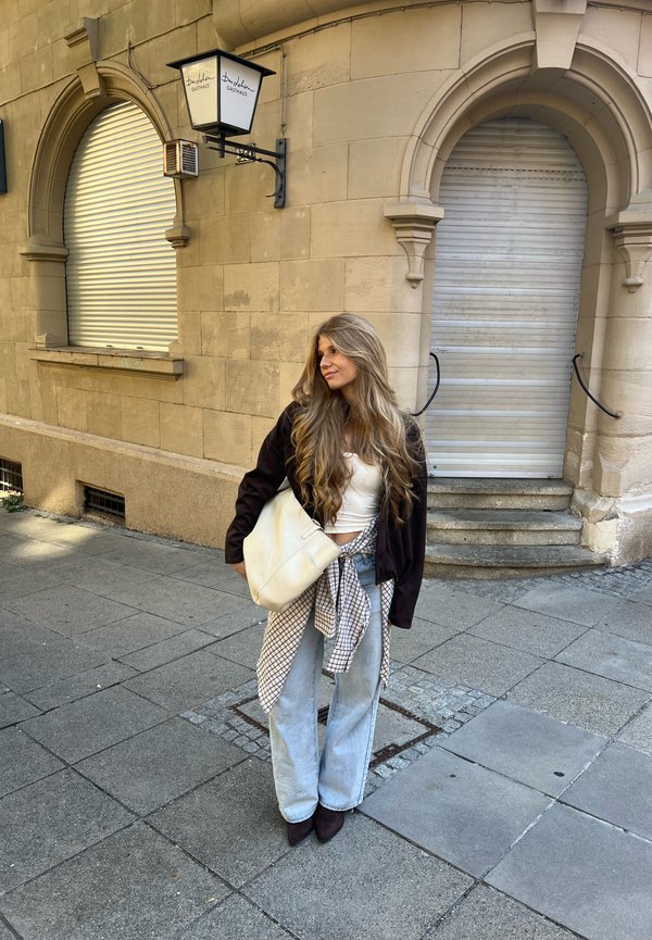 Mujer con el pelo largo está de pie en una acera frente a un edificio de piedra beige con contraventanas arqueadas cerradas, viste vaqueros claros y una chaqueta oscura.