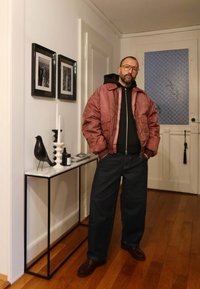 Gaiši burgundas bomberjaka, melna kapuce, tumši zili plataji džinsi, brūnas kurpes. Balts konsoles galds ar dekoratīviem priekšmetiem un rāmētiem fotoattēliem.