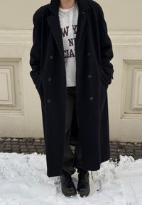 Long manteau en laine noir, sweat-shirt gris avec le texte "NEW YORK", pantalon noir et bottes noires à lacets. Neige et pavés visibles en dessous.