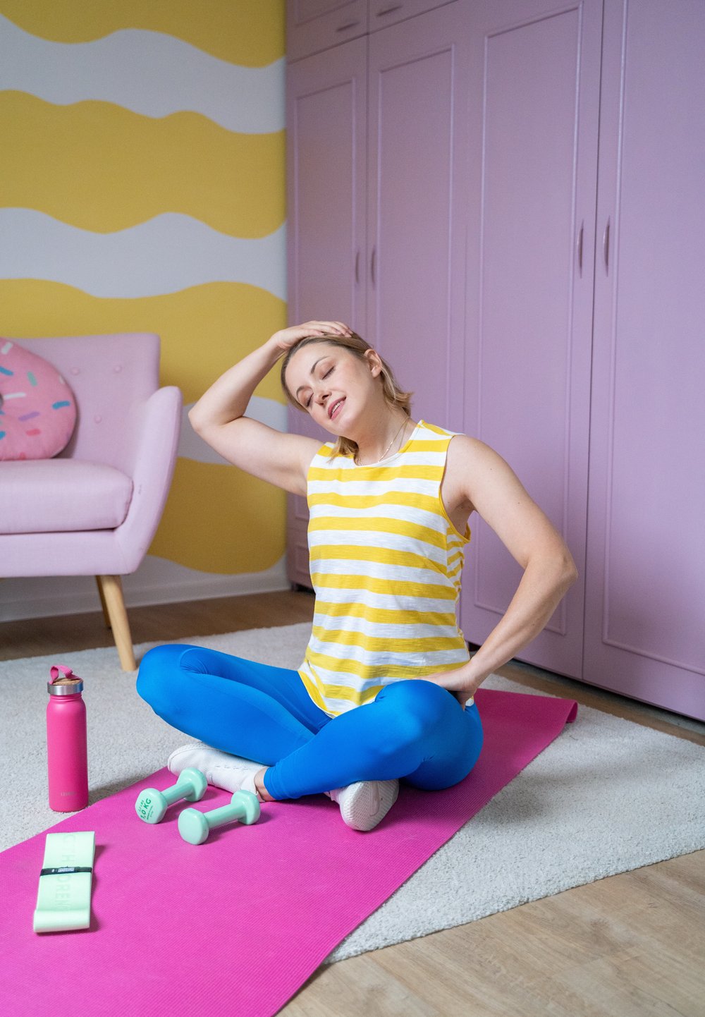 Αθλητής σε κίτρινο ριγέ κάνει τοπ και μπλε κολάν, καθισμένος σε ροζ στρώμα με αλτήρες και μπουκάλι νερού κοντά. Ροζ τοίχοι και χαλί.