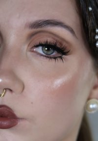 Primo piano di un volto che mette in evidenza un occhio verde con lunghe ciglia, ombretto bronzo e un anello al setto nasale dorato. Pelle morbida e luminosa e labbra opache di un colore intenso.