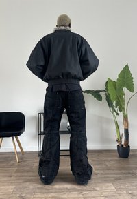 Czarna kurtka bomberka z puszystym kołnierzem, w połączeniu z czarnymi, postrzępionymi spodniami cargo z widocznym przeszyciem. Wnętrze z roślinnością.