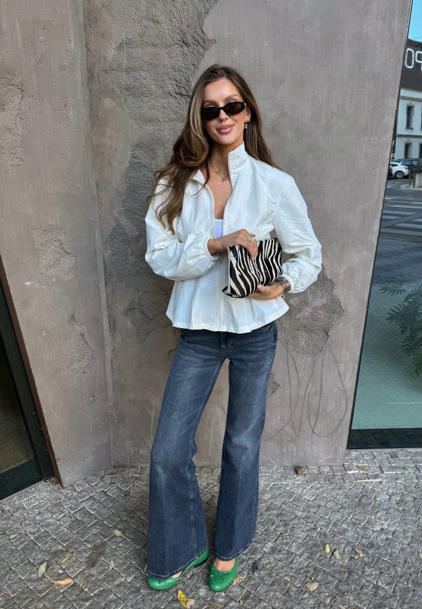 Femme portant une veste blanche, un jean bleu, des chaussures vertes et des lunettes de soleil noires, tenant une pochette à motif zèbre, debout contre un mur texturé.