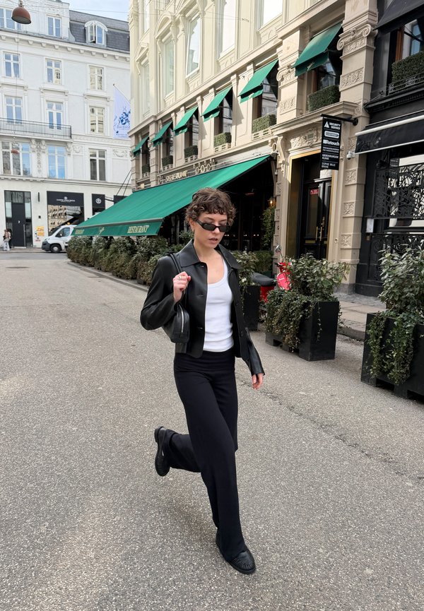 Jeune femme portant une veste en cuir noire, un haut blanc et des lunettes de soleil, marchant dans une rue de la ville avec des auvents de restaurant verts et des plantes en pot en arrière-plan.