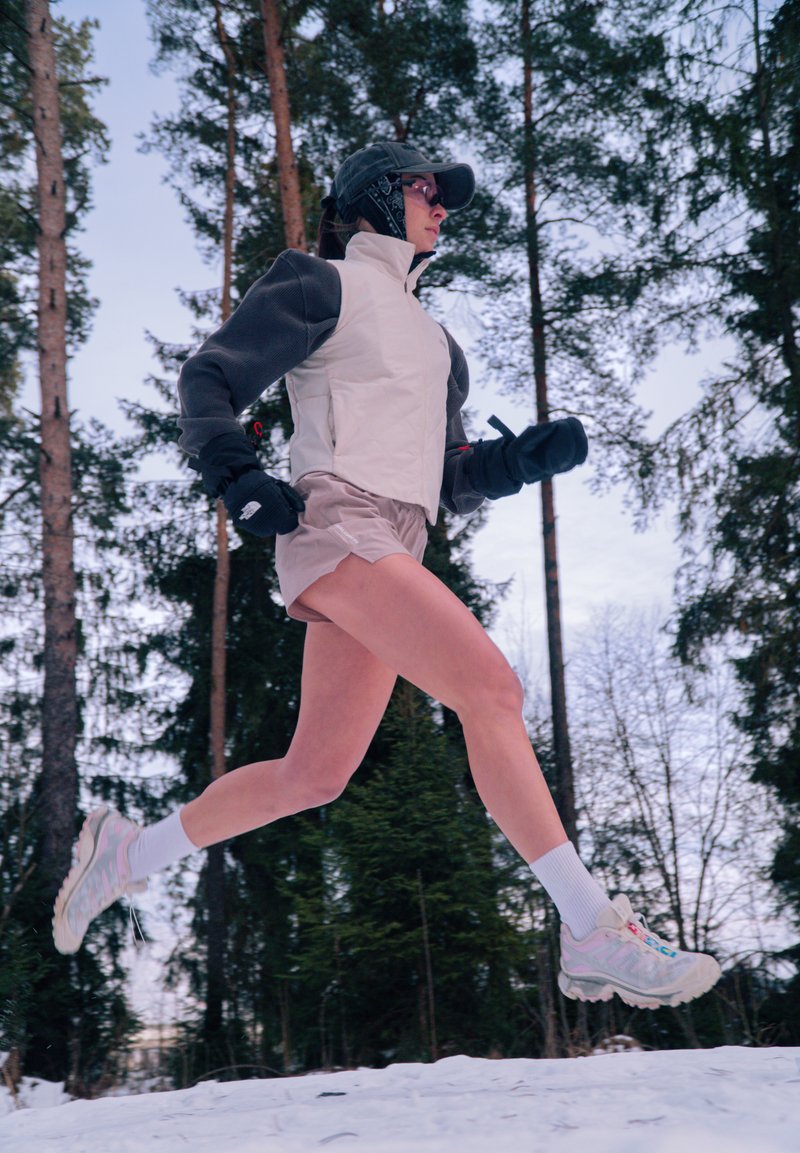 Personne portant une casquette, une veste, un short et des gants en plein saut en courant sur un sentier enneigé dans une forêt de grands pins.