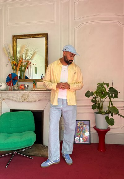 Hombre con camisa amarillo claro, camiseta blanca, gorra azul y zapatillas deportivas de pie en una habitación con silla verde, planta sobre un taburete rojo y arte enmarcado en el suelo.