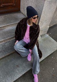 Casaco de pele sintética castanho, cardigan rosa, jeans largos cinzentos e ténis rosas. Os acessórios incluem um gorro preto canelado e óculos de sol.