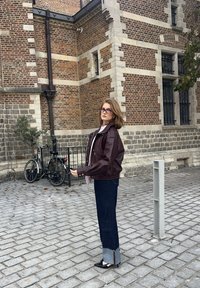 Vinröd läderjacka, ljusrosa krage, mörka denimjeans med rå fåll, svarta spetsiga skor; i bakgrunden syns en tegelvägg och cyklar.