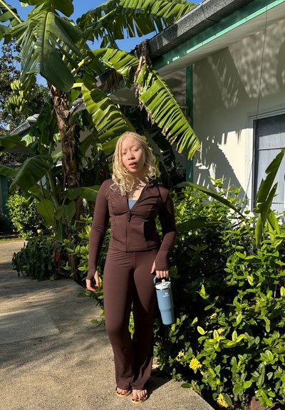 Conjunto deportivo marrón con chaqueta con cremallera y pantalones ajustados. Sosteniendo una botella de agua azul claro. Fondo con vegetación y luz solar.