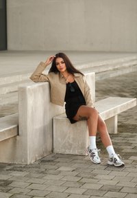 Beige kort jacka över en svart topp och kjol, vita sportstrumpor och mångfärgade sneakers. Sitter på en betongbänk.