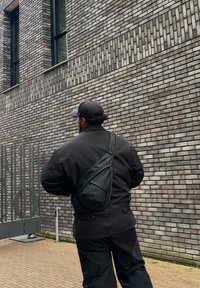 Black sling bag made from synthetic material, featuring a curved shape and smooth texture, worn over a black jacket against a brick wall.
