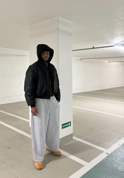 Chaqueta bomber negra, pantalones de chándal grises y botas beige. La persona está de pie en un garaje de estacionamiento con paredes blancas y un letrero de flecha verde.
