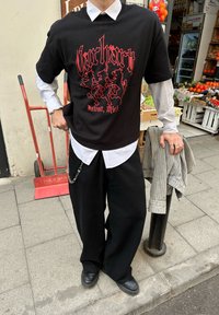 T-shirt noir surdimensionné avec texte graphique rouge, superposé sur une chemise blanche à col. Pantalon noir ample, détail chaîne, et chaussures noires.