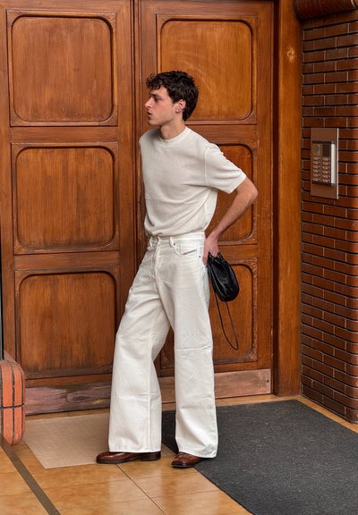 Vaqueros blancos de pierna ancha, camisa de punto crema texturizada, bolso negro y zapatos marrones. De pie junto a puertas de madera, mostrando un estilo casual.