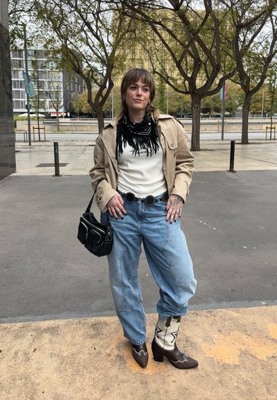 Gabardina beige, suéter blanco de canalé, bufanda negra con flecos, jeans azules de gran tamaño, cinturón negro con acentos circulares, bolsa negra, botas de cowboy con detalles blancos.