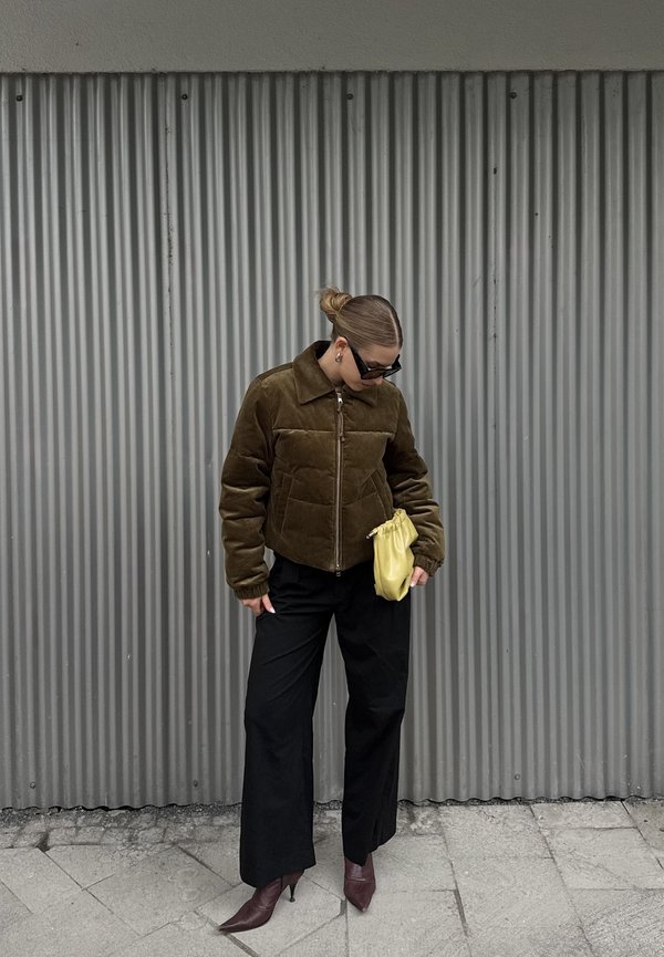 Braune, gekürzte, gesteppte Jacke mit Kragen, getragen über schwarzen weiten Hosen. Hält eine kleine, gelbe Tasche und trägt eine Sonnenbrille.