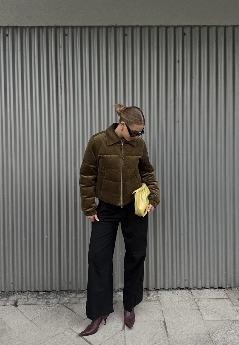 Veste brune, courte et matelassée avec un col, portée sur un pantalon noir à jambes larges. Tient un petit sac pochette jaune et porte des lunettes de soleil.