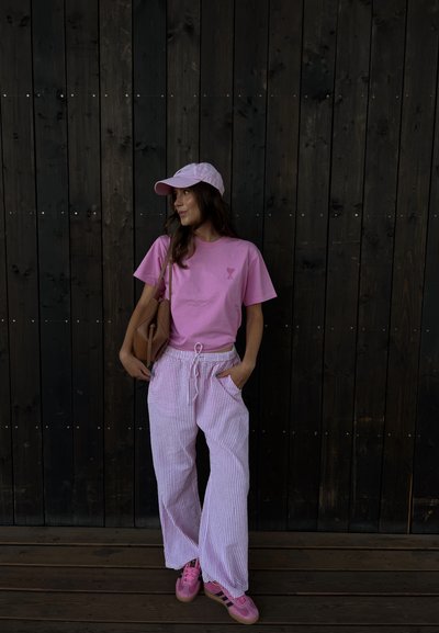 Camiseta rosa con un pequeño gráfico, pantalones de pierna ancha a rayas en colores claros, zapatillas rosas y una gorra a juego. Bolso marrón colgado del hombro.