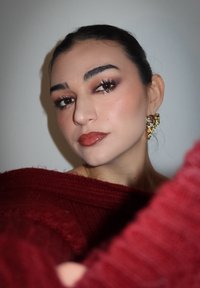 Portrett med en mørkerød strikkegenser, med dristige øredobber. Ansiktet har varme sminketoner med definerte øyne og glansfulle lepper.