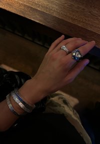 Ezüst karkötők texturált mintákkal, egy vékony gyűrű és egy nagy, szív alakú gyűrű óraszámlappal egy kézen. Sötét háttér.