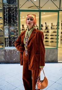 Completo marrone in velluto a coste con blazer oversize, camicia verde con colletto, cravatta fantasia e borsa arancione con accessori portachiavi colorati.